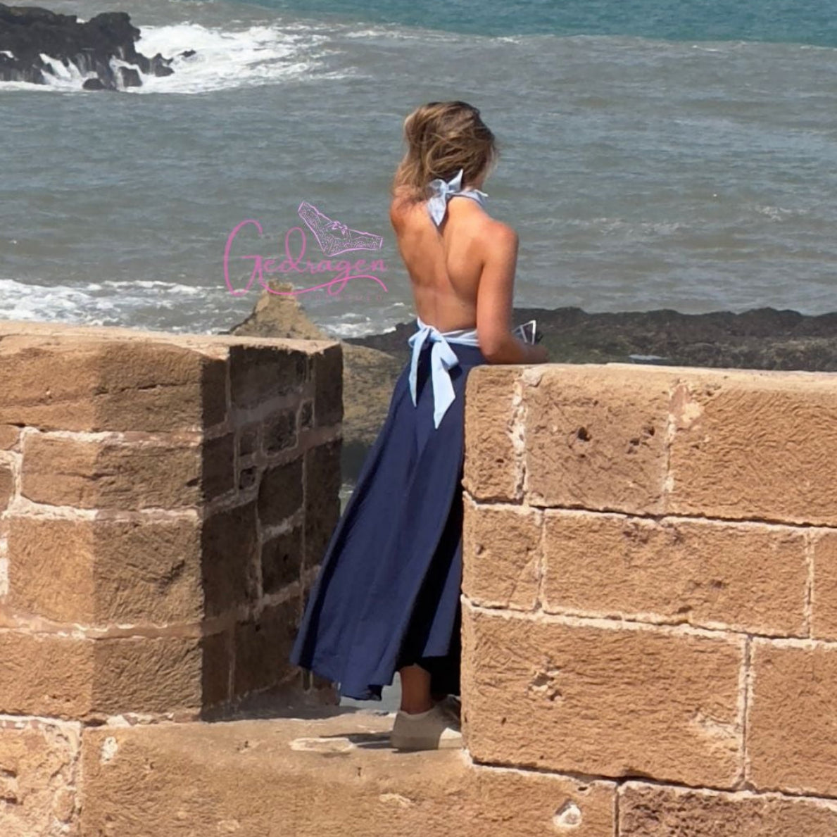 Vrouw in een elegante rugloze blauwe jurk kijkt uit over de zee vanaf een stenen uitkijkpunt, romantische sfeer aan de kust.